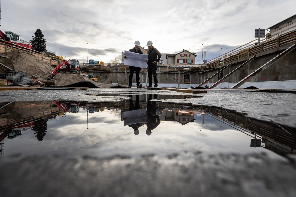 Man sieht zwei Männer, die in einer Baugrube einen Plan studieren. Sie spiegeln sich in einer Pfütze am Grund.
